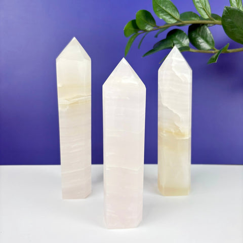 Three polished pink calcite crystal towers with banded textures standing on a white surface against a purple background.
