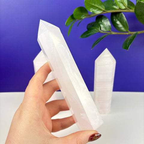 Hand holding a translucent, pale pink calcite crystal tower against a blue background with two other towers nearby.