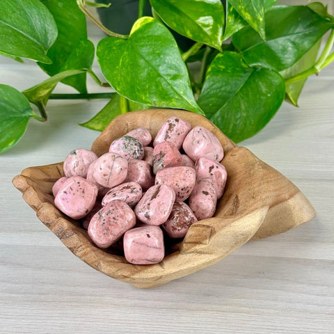 Pink Cobalto Calcite tumble stones in a natural wood bowl, with green plant leaves in the background.