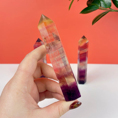Hand holding a polished Poison Candy fluorite crystal tower with vibrant purple, yellow, and red banded layers.
