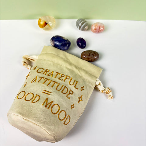 Cream cotton-linen drawstring pouch with gold embroidered text and ribbon, surrounded by assorted polished crystals.