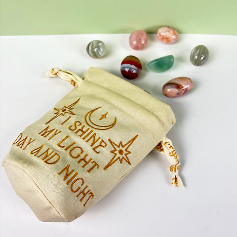 Cream-colored canvas drawstring pouch with gold embroidery, ruffled top, and assorted tumbled crystals.