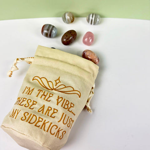 Cream canvas drawstring pouch with gold embroidery and ruffled top, surrounded by polished crystals.