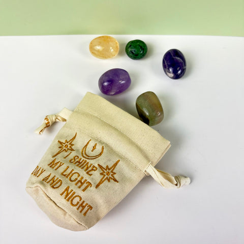 Small beige cotton-linen drawstring pouch with gold embroidery and five polished crystals on a white surface.