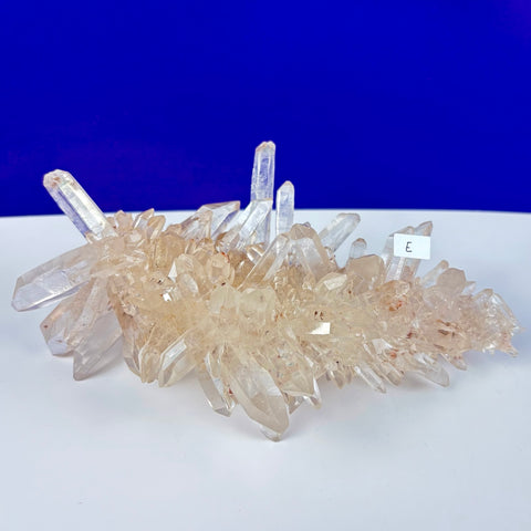 Cluster of clear Himalayan quartz crystals with reddish inclusions on a white surface against a blue background.