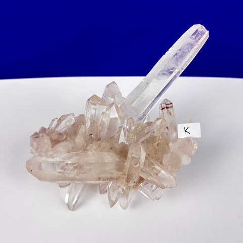 Clear Himalayan quartz crystal cluster with a prominent, elongated central point on a white surface.