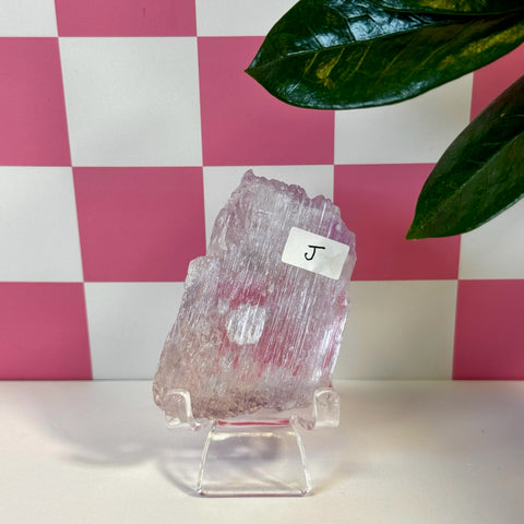Vibrant pink Kunzite crystal, raw and translucent, displayed on a clear stand with a checkered background.
