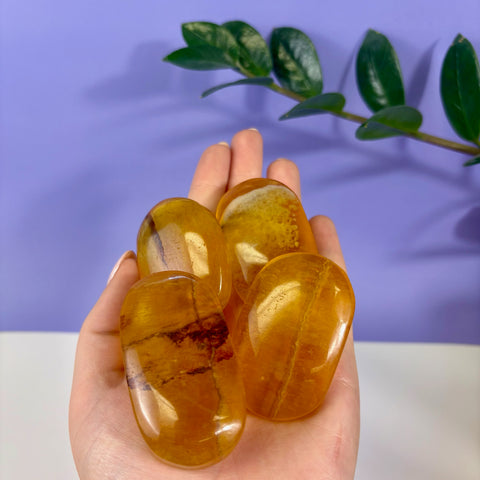 Four polished, translucent yellow fluorite palm stones with natural inclusions held in a hand against a purple background.
