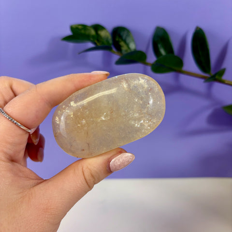 Polished yellow fluorite palm stone held in hand against a purple background with green foliage.