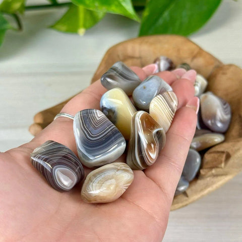 Polished grey, white, and brown banded agate tumble stones held in a hand, with more in a wooden bowl.