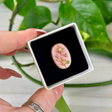 Pink oval enamel pin with gold stars and crystal accents in a white box.
