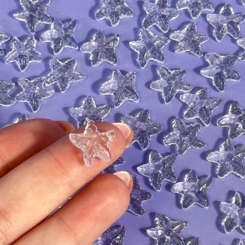 Mini clear quartz sea star held between two fingers, with many more scattered on a purple surface.