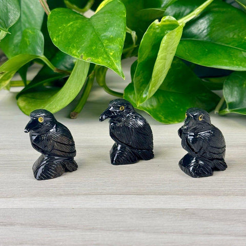 Three dark, glossy raven figurines with yellow eyes, set against a light wood surface and green leaves.