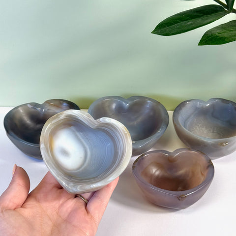Hand holding a polished gray and white agate heart bowl, with four similar bowls in the background.