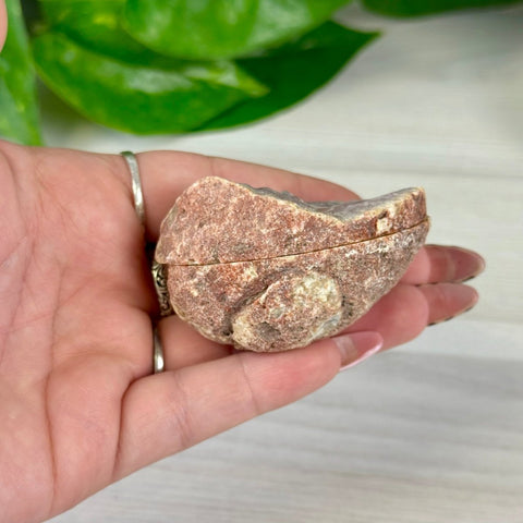 Hand holding a small, rough-hewn reddish-brown agate geode pair, partially open to reveal crystals.