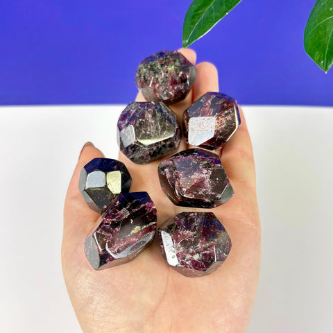 Hand holding six polished, deep red Cherry Blood Garnet crystals with faceted geometric shapes.