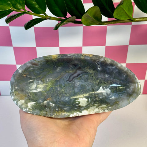 Curvy moss agate bowl with green, white, and gray patterns, held against a pink and white checkered background.