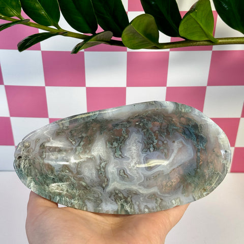 Curvy moss agate bowl with natural green, white, and brown swirls, held against a pink and white checkered background.
