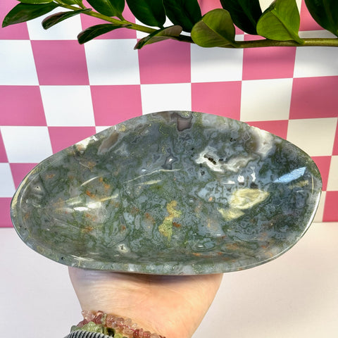 Curvy moss agate bowl with natural green, white, and brown patterns, held against a pink and white checkered background.