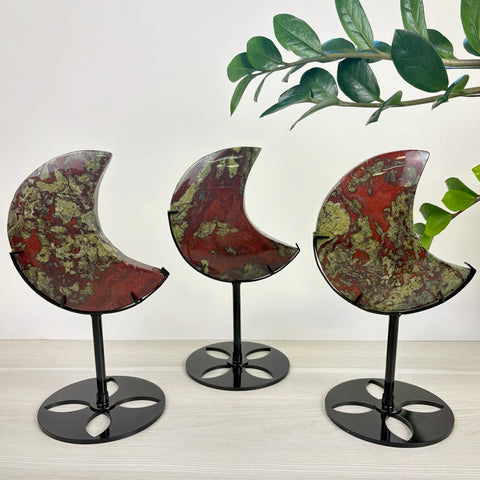 Three crescent moon-shaped Dragon's Blood jasper stones with green and red patterns displayed on black metal stands.