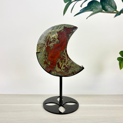 Polished crescent moon-shaped Dragon's Blood stone with red and green patterns, displayed on a black metal stand.