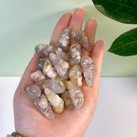 Hand holding multiple polished flower agate spiral shell carvings in varying translucent pink, white, and cream tones.