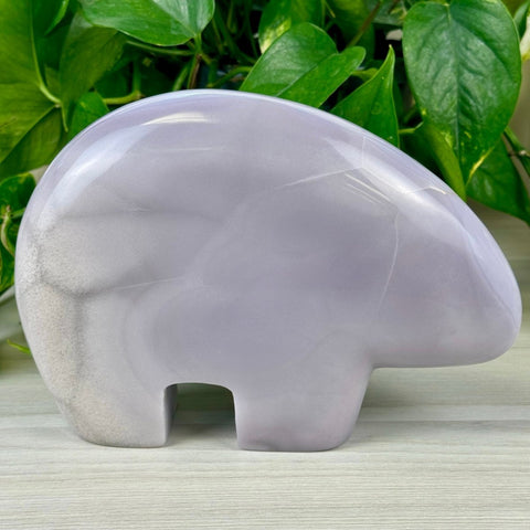 Gabby The Bear carving, a smooth lavender fluorite crystal with subtle white veins, against green leaves.