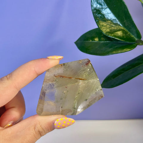 Hand holding a clear garden quartz free form with green and red inclusions.