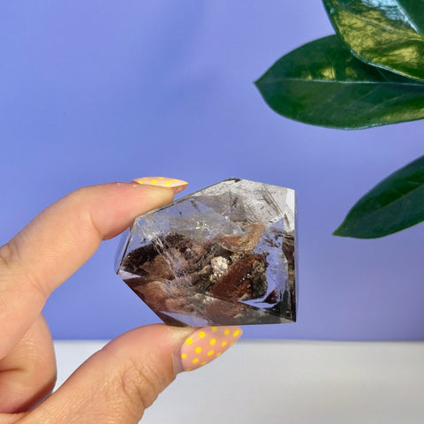 Hand holding a clear garden quartz free form with dark brown and red inclusions.