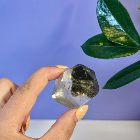 Hand holding a clear Garden Quartz Free Form with dark green inclusions, against a purple background.