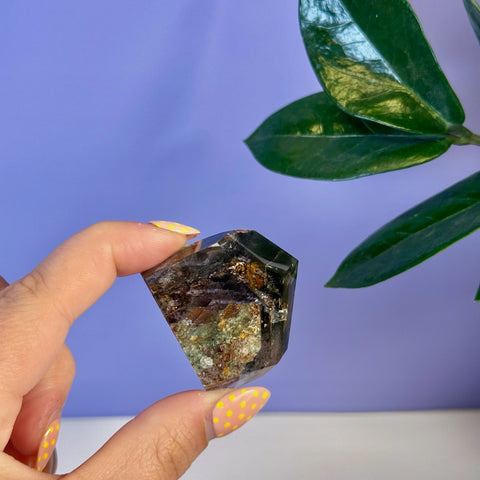 Hand holding a dark, multifaceted Garden Quartz free form with green, brown, and purple inclusions.