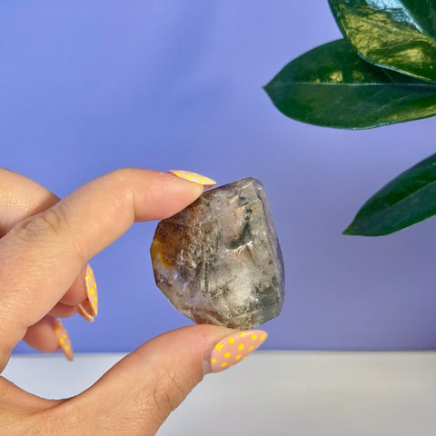 Hand holding a small, clear garden quartz free form with brown and black inclusions.