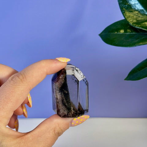 Hand holding a dark, faceted garden quartz free form with visible inclusions, against a purple background.
