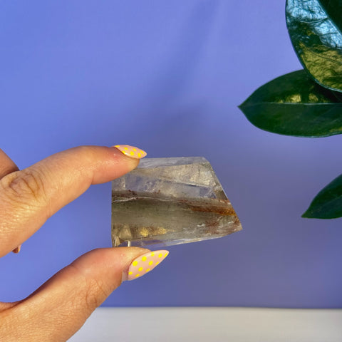 Hand holding a clear garden quartz free form with green and reddish-brown inclusions.