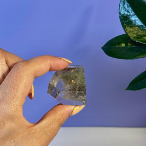 Hand holding a clear garden quartz free form with green inclusions against a purple background.