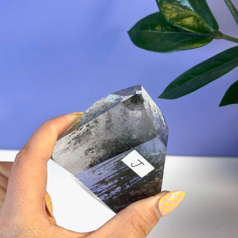 Hand holding a clear garden quartz free form with dark inclusions and a "J" sticker.