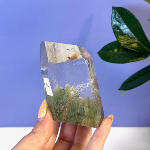 Clear garden quartz free form with green chlorite inclusions, held against a purple background.