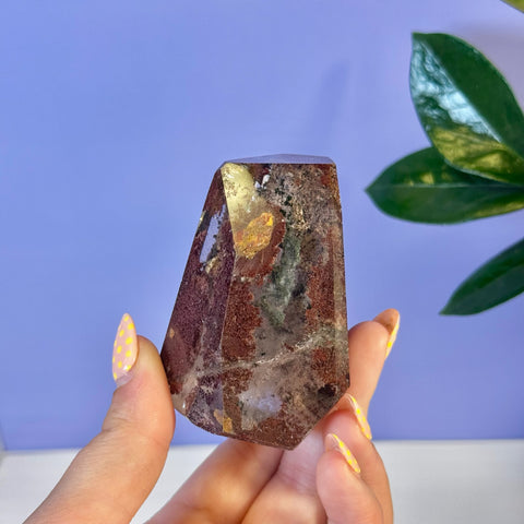 Hand holding a dark red and clear Garden Quartz free form with golden and green inclusions.