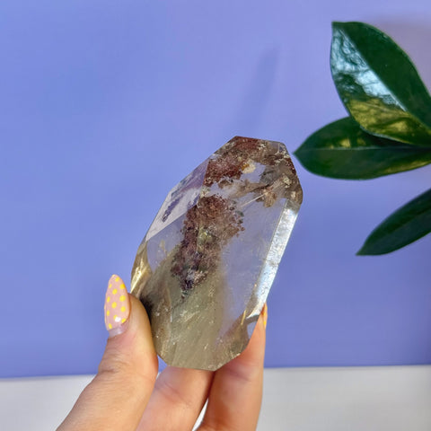 Clear garden quartz free form crystal with reddish-brown inclusions, held by a hand with yellow polka dot nails.