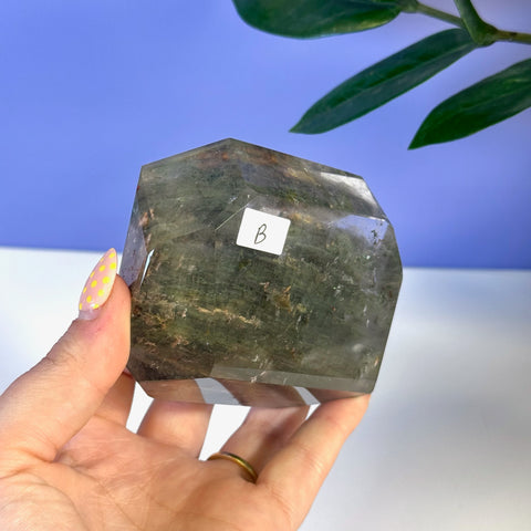 Hand holding a faceted, dark green Garden Quartz free form with natural inclusions.