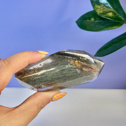 Hand holding a clear Garden Quartz free form with green, red, and gold inclusions.