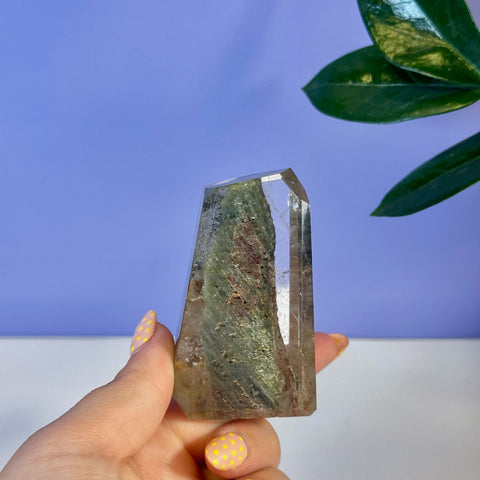 Clear garden quartz free form with green and reddish inclusions, held in hand.