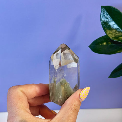 Hand holding a clear garden quartz free form crystal with green inclusions and a faceted top.