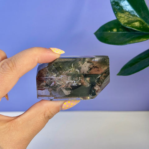 Hand holding a clear Garden Quartz free form with green, red, and brown mineral inclusions.