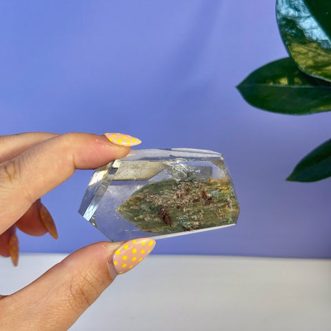 Hand holding a clear, faceted Garden Quartz Free Form with green and reddish inclusions.