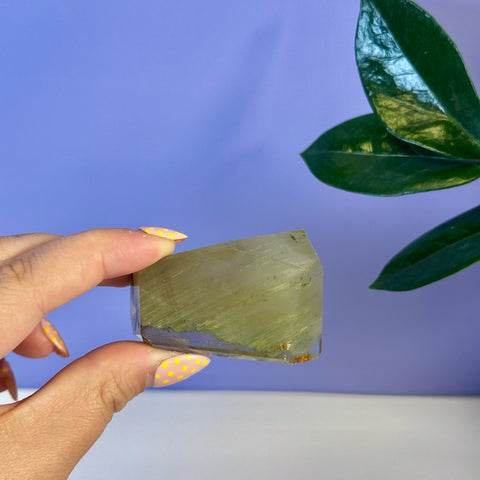 Hand holding a clear Garden Quartz Free Form with green inclusions, against a purple background.