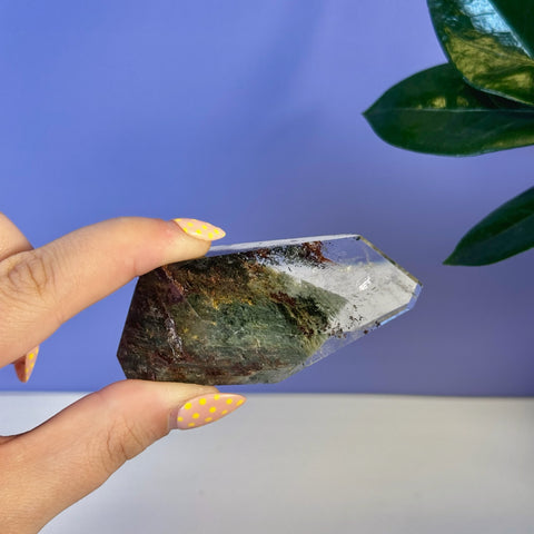 Hand holding a clear garden quartz free form with green and reddish-brown inclusions.