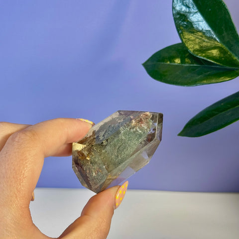 Hand holding a clear Garden Quartz free form crystal with green, red, and brown inclusions.