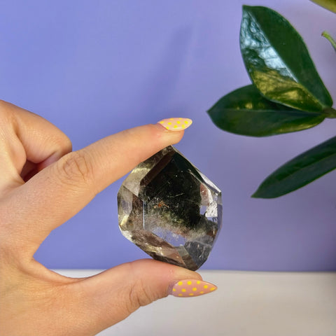 Hand holding a clear garden quartz free form crystal with dark green inclusions.