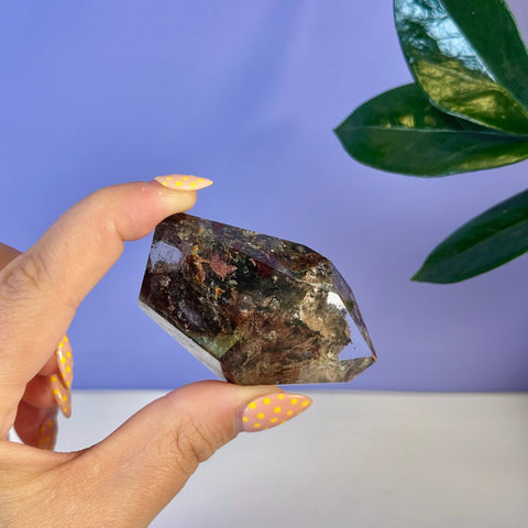 Garden Quartz free form crystal, clear with dark red and green mineral inclusions, held by fingers with yellow polka dot nails.